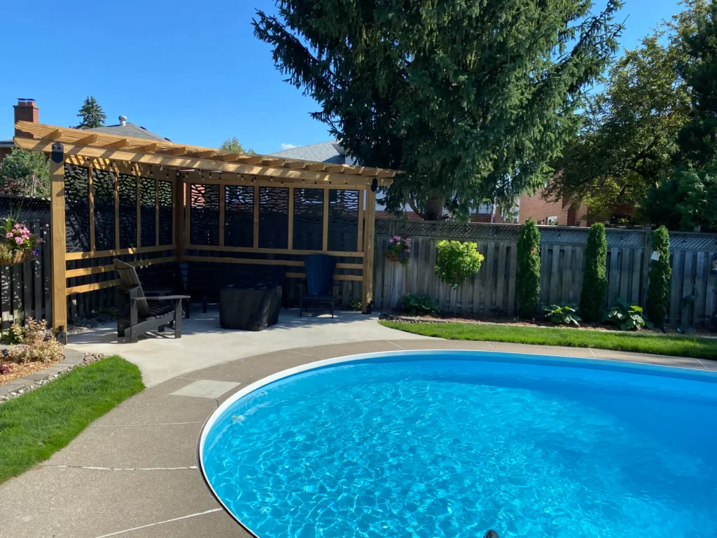 backyard landscaping with swimming pool, wood pergola seating area, concrete patio, and green lawn with trees and fence