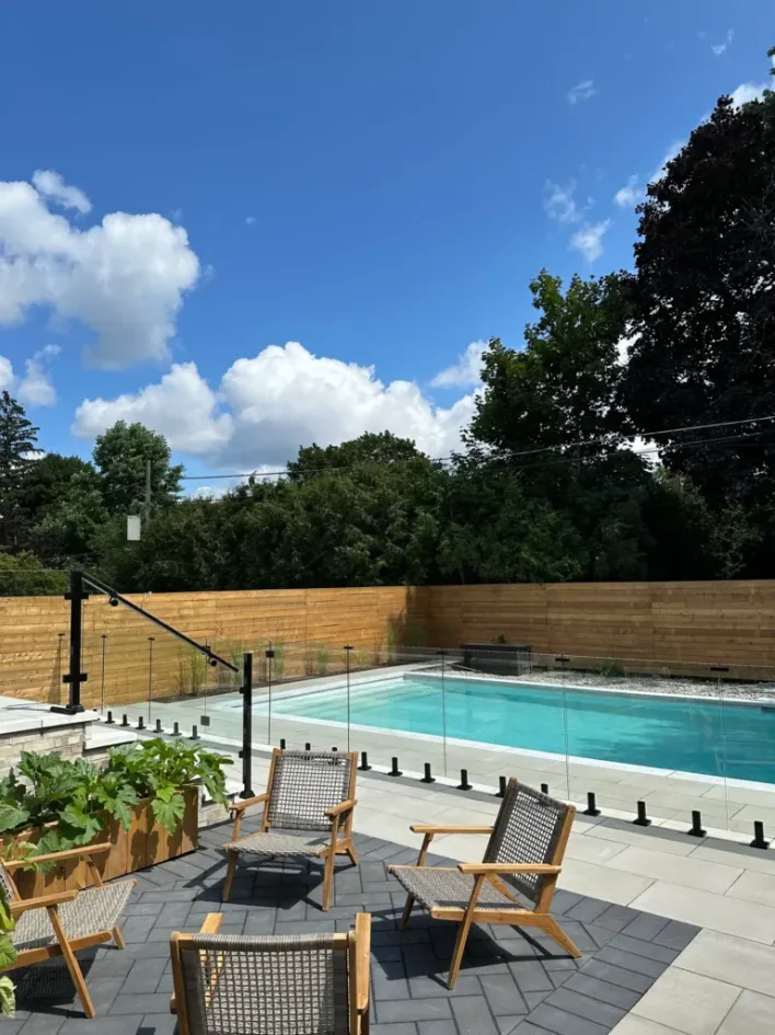 modern backyard with rectangular pool, glass railing, stone patio, wooden fence, and outdoor lounge chairs under blue sky