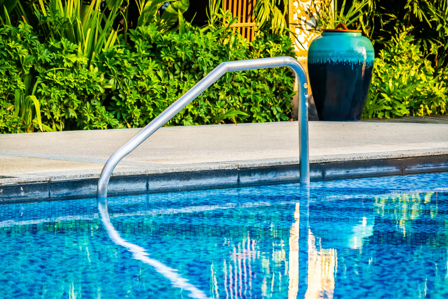 swimming pool with metal handrail, blue water, tropical plants, and decorative ceramic pot in landscaped garden