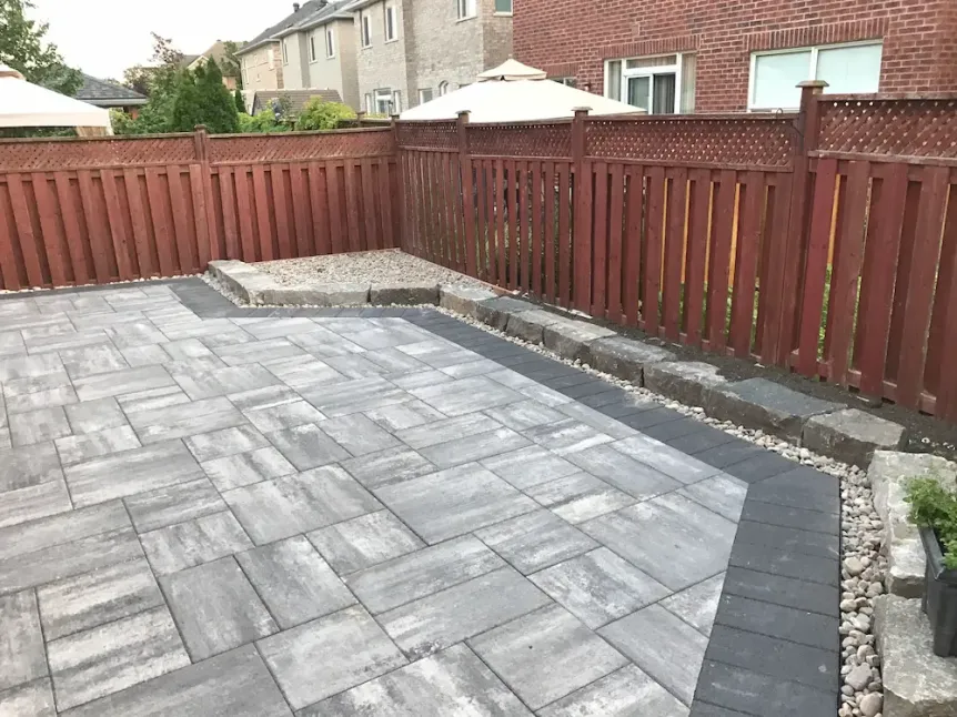 Modern backyard patio with gray interlocking pavers, dark border accent, stone edging, and red wooden privacy fence