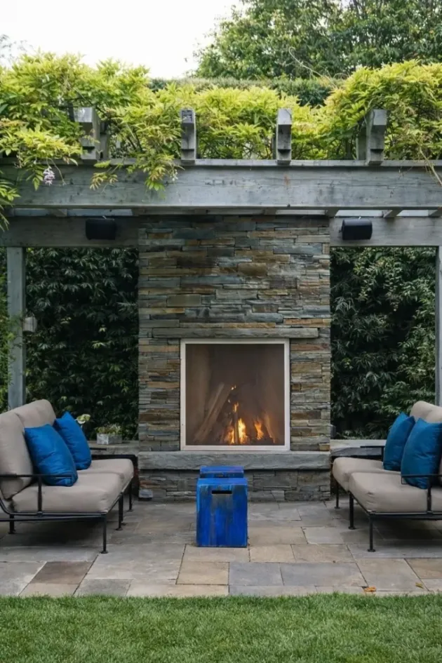 modern outdoor fireplace with stone wall, pergola overhead and cushioned seating area