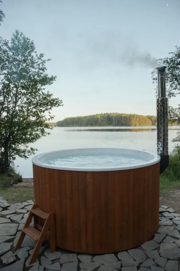 Round wood-fired hot tub with a chimney heater beside a calm lake at dusk, photo by oakshs.ca