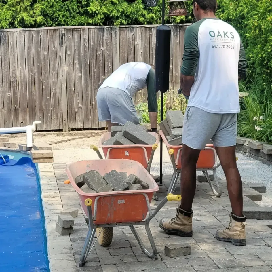 workers installing interlocking pavers near pool patio with stone blocks and wheelbarrows
