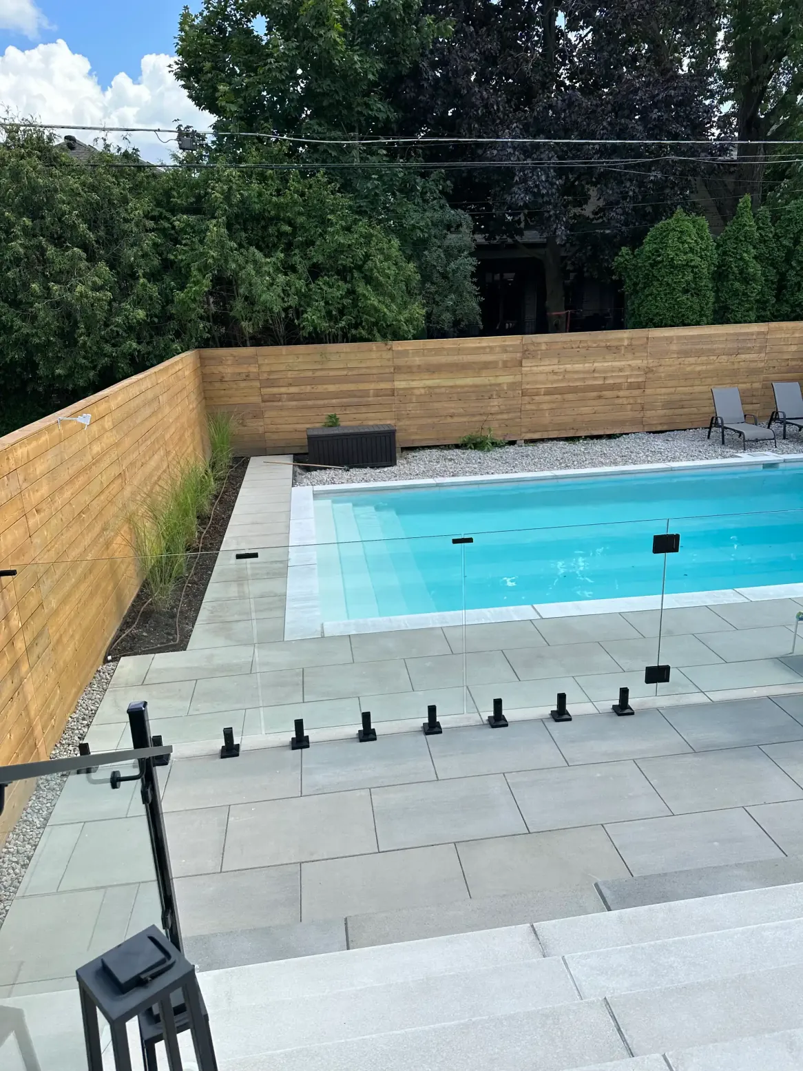 modern backyard with swimming pool, glass fence railing, large stone patio, wood privacy fence, and clean minimalist landscaping