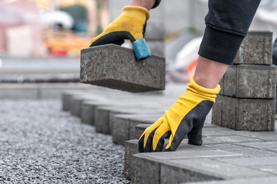 Contractor installing interlocking pavers on gravel base with protective gloves