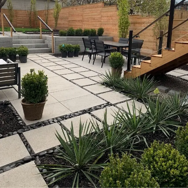 Backyard patio with concrete pavers, decorative gravel joints, wood stairs, and landscaped garden beds, image by OAKS Home Services
