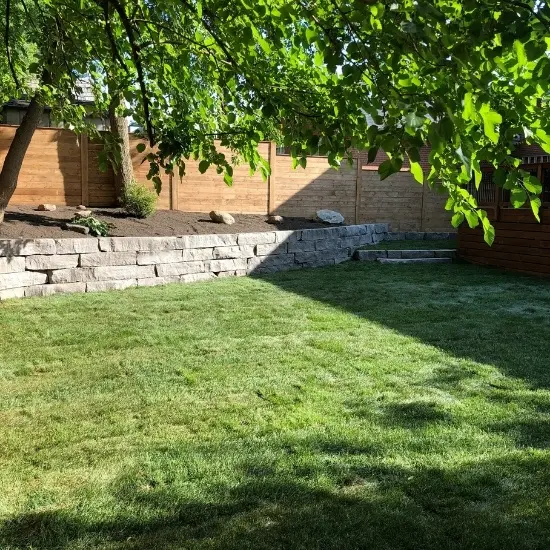 Backyard landscaping with stone retaining wall, lawn, and wooden privacy fence, image by OAKS Home Services