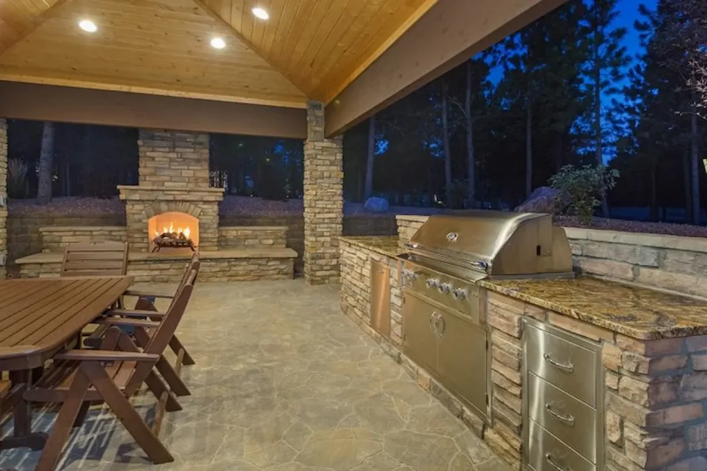 Covered outdoor kitchen with built-in grill, stone fireplace, and dining area, image by OAKS Home Services