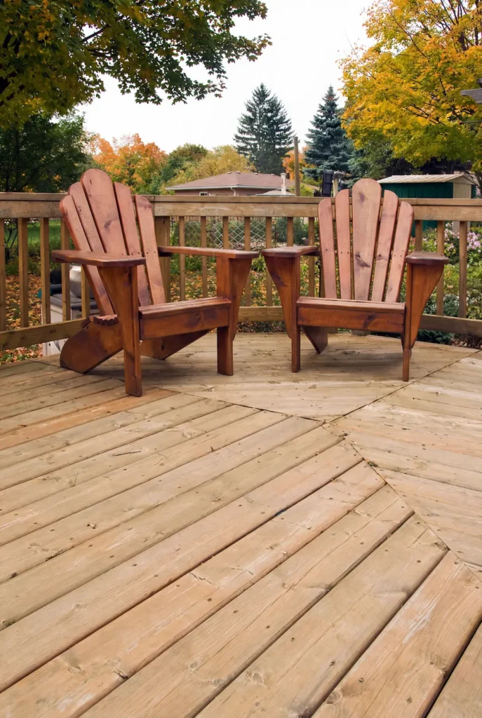 Two wooden Adirondack chairs on a backyard deck surrounded by autumn trees